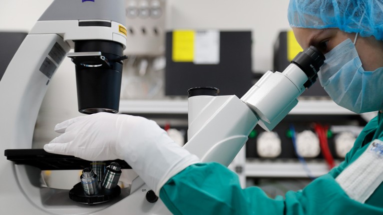 A researcher in mask, gloves and hairnet looks through a microscope.