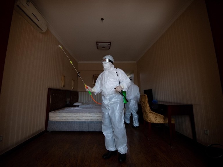 A person in full protective gear sprays a room containing a bed, desk and chair.