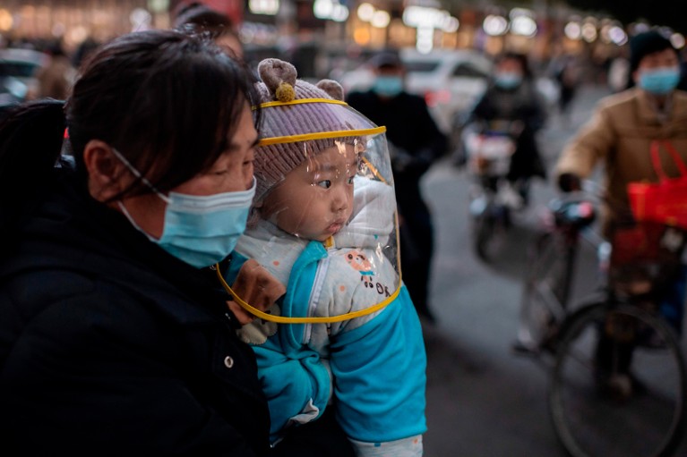 A woman wearing a face mask holds a baby wearing a protective shield.
