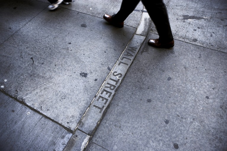 The shoes and legs of a businessman are seen walking over a sign on the street saying "Wall Street"