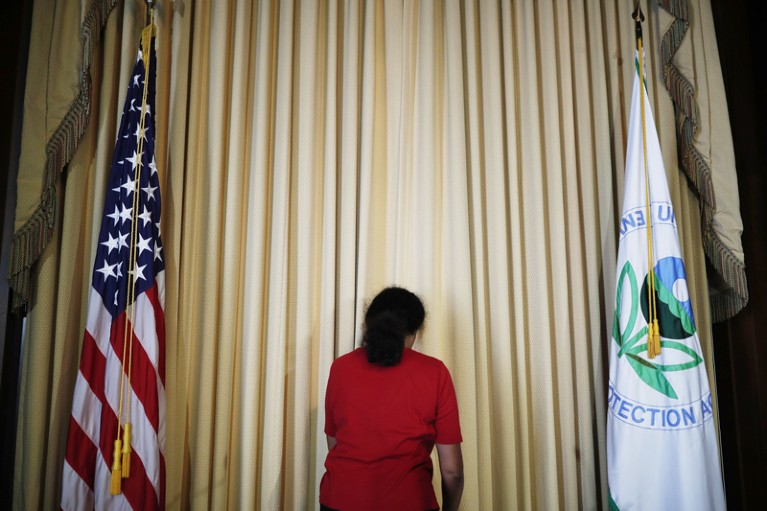 A woman flanked by flags of the United States and the US Environmental Protection Agency smooths curtains on a stage.
