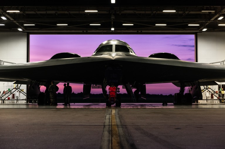 A straight-on view of a B-2 stealth aircraft inside a hanger with a purple sky seen through the door