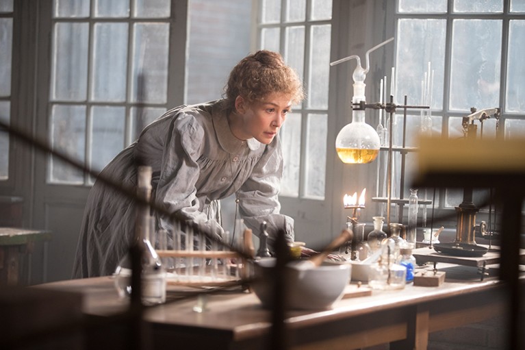 A woman in a loose Edwardian dress leans over a lab bench.