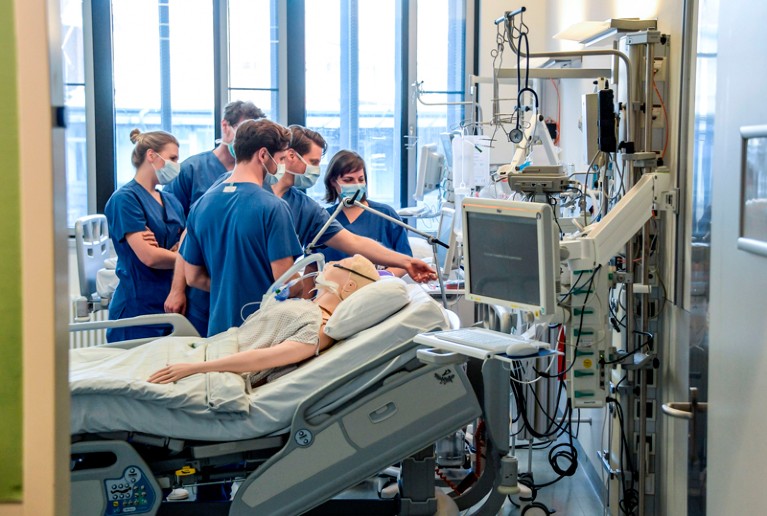 A group of doctors in scrubs and face masks are instructed on how to use a ventilator on a dummy