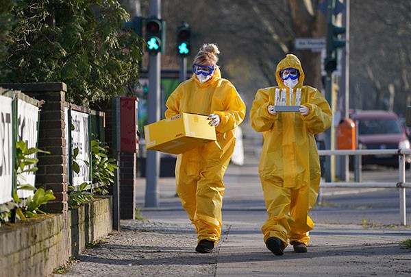 A pair of medical volunteers in full protective outfits who hold samples while walking down a street