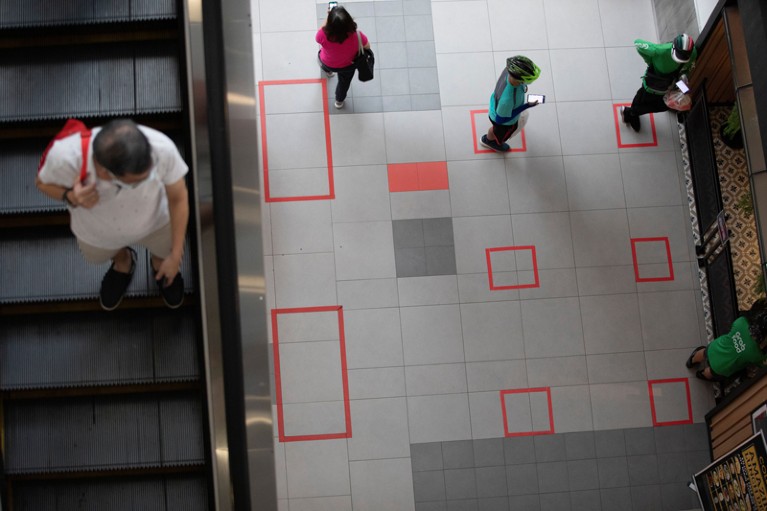 A man on an escalator looks down at people standing in squares marked on the floor as part of social distancing measures.