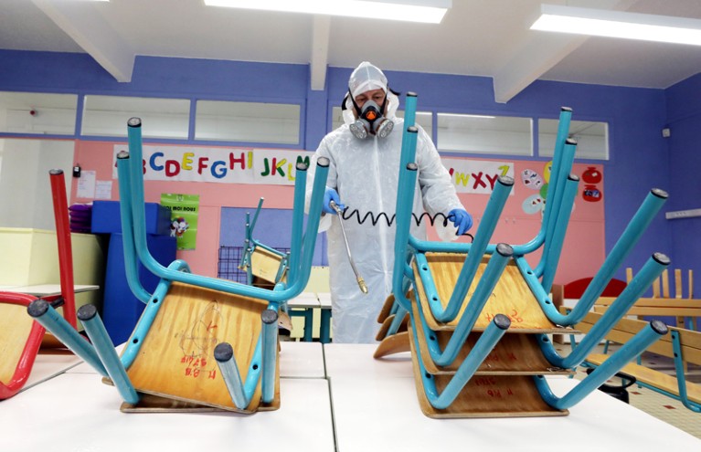 Worker in a protective suit and respirator mask sprays disinfectant on chairs and desks in a classroom
