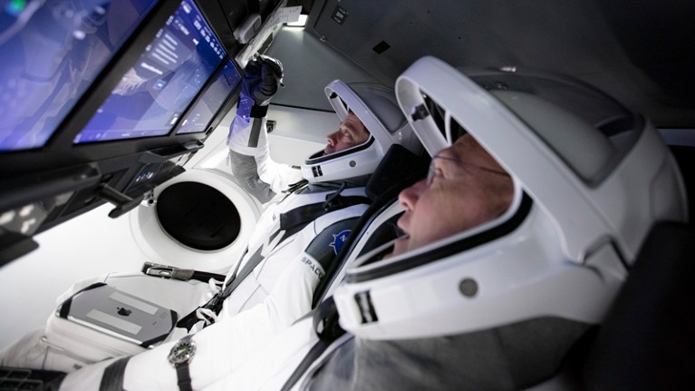 Astronauts Bob Behnken and Doug Hurley wearing space suits and helmets touch control panels during a flight simulation