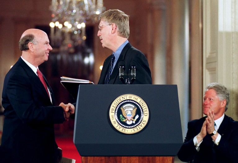 Francis Collins and Craig Venter shake hands at a podium while Bill Clinton looks on and applauds during a press conference
