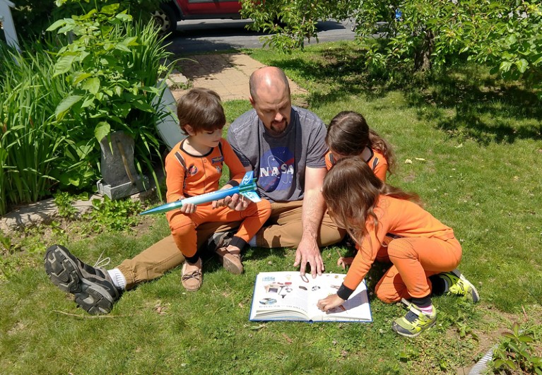 Jeremy Grabbe reads from a book while sitting on the lawn with his triplets who are wearing matching spacesuit outfits