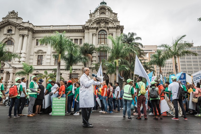 Salim Abdool Karim, wearing a white coat and holding a megaphone, leads a crowd of demonstrators in Durban, South Africa