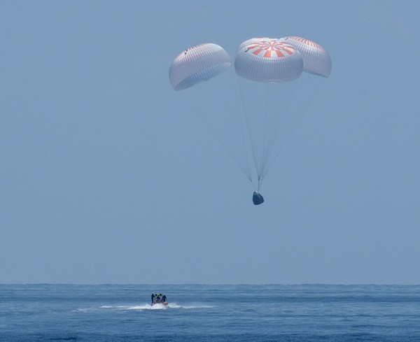 A small motorboat approaches a spacecraft attached to four parachutes about to land on the Gulf of Mexico.