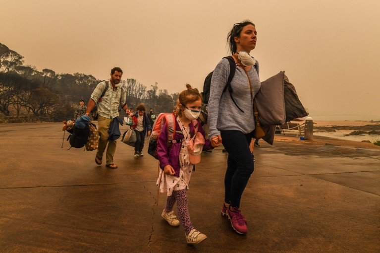 A family carrying personal belongings walk under smoke-filled skies as they are evacuated away from a wildfire