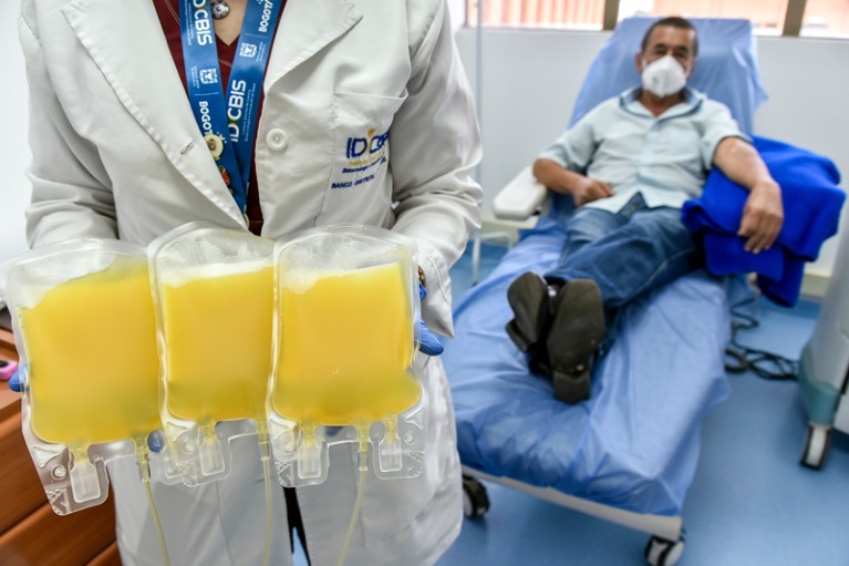 A health worker holds three bags of donated plasma while a donor in a face mask sits on a hospital bed.