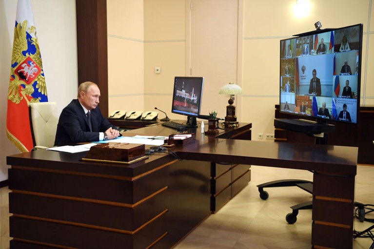 Vladimir Putin sitting at a large desk, watching a videoconference, with a flag behind him.