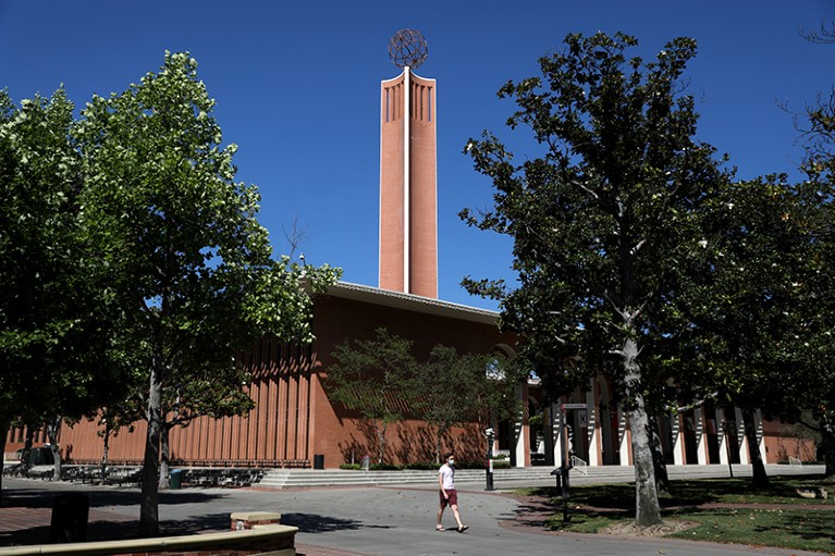 A brown building with a tall central tower, seen through trees against a bright blue sky.