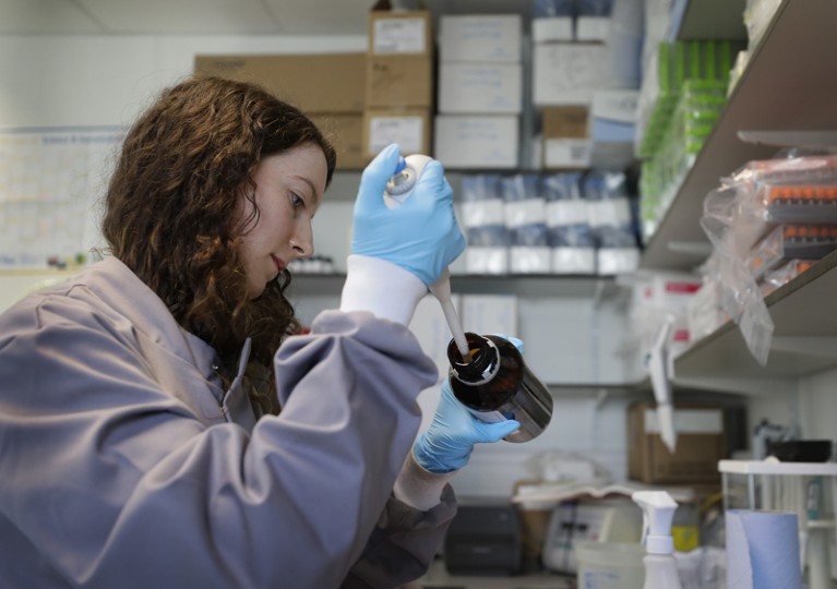 A woman wearing a lab coat and blue gloves pipetting liquid from a bottle in a laboratory