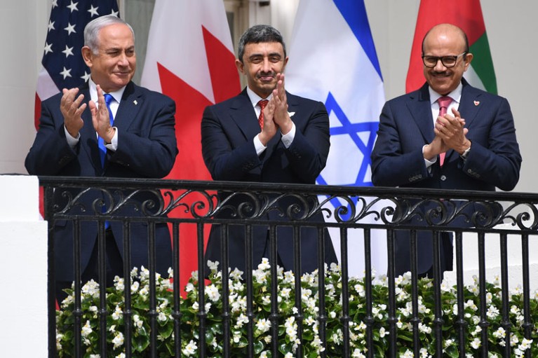 Three men in suits applauding on a balcony, standing in front of the flags of the United States, Bahrain, Israel and the UAE.