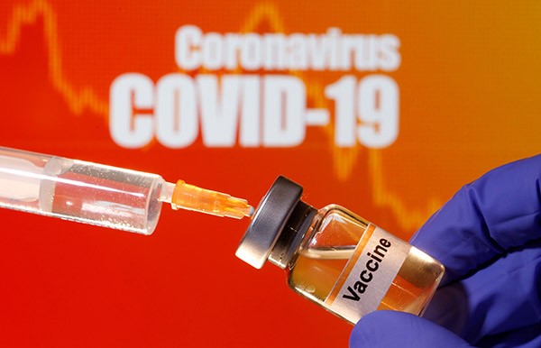A small bottle labeled with a "Vaccine" sticker is held near a medical syringe in front of "Coronavirus COVID-19" signage