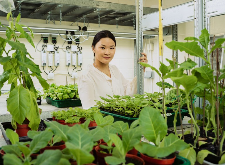 Audrey Teh poses for a portrait in an artificially lit room surrounded by seedlings growing in pots
