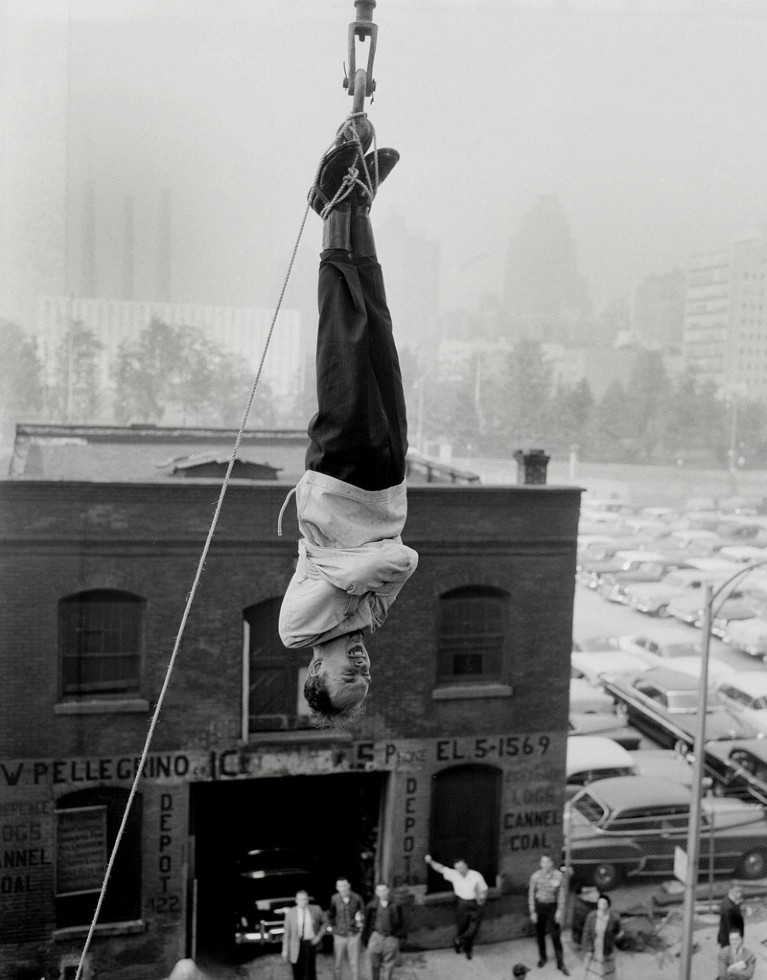 James Randi performing an escape stunt in a strait jacket while suspended from a crane above New York in 1956