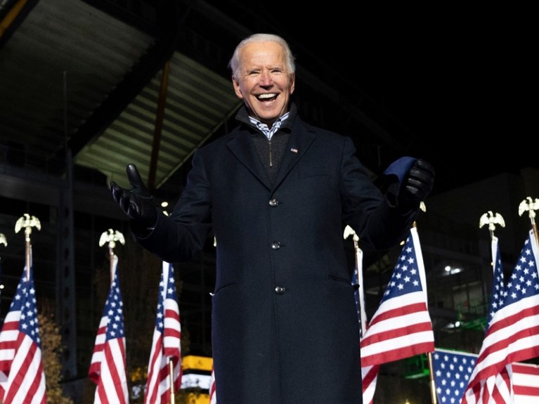 Joe Biden smiling in front of a row of US flags.