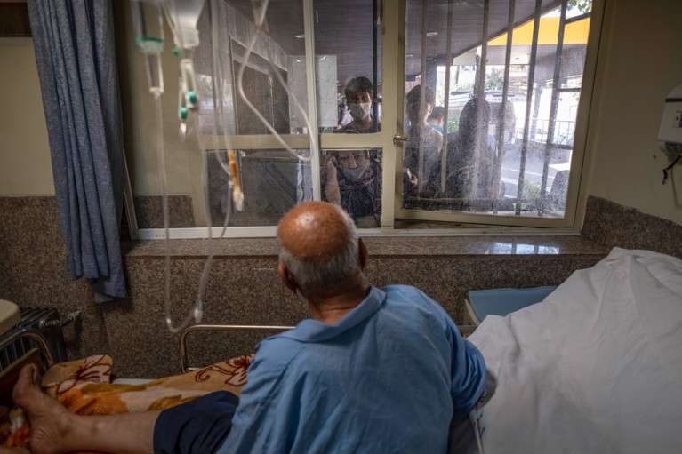 An elderly male patient in a hospital bed talks to relatives through a window in a hospital in Iran