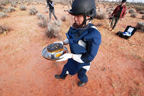 A person wearing heavy duty protective equipment in the desert carries a dinner-plate-sized capsule.