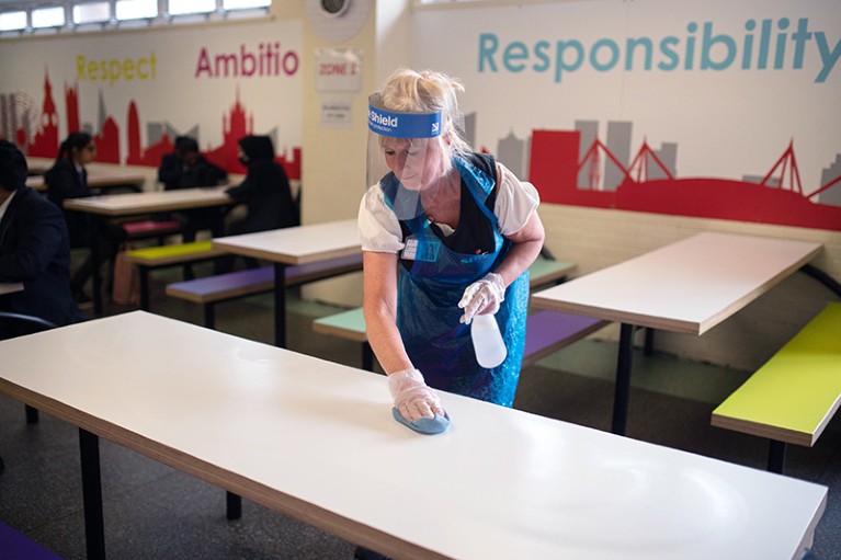 A cleaning staff member wearing PPE sanitises tables and chairs in a school
