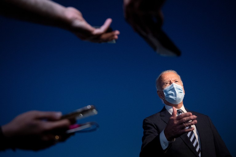 Joe Biden wearing a face mask talks to reporters pointing microphones at him