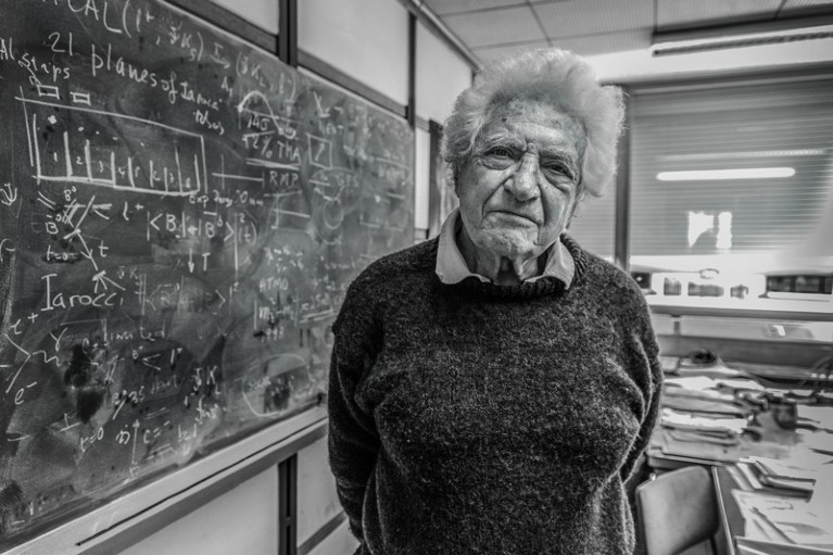A black and white portrait of Jack Steinberger in his office standing by a large blackboard covered in chalk writing