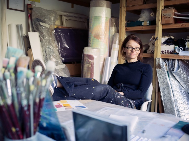 Geraldine Cox poses for a portrait in her artists studio with her feet up on the desk, surrounded by brushes and canvases