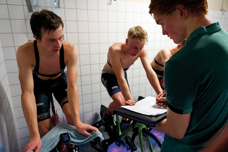 A scientist records data while standing in front of male athletes on static bikes