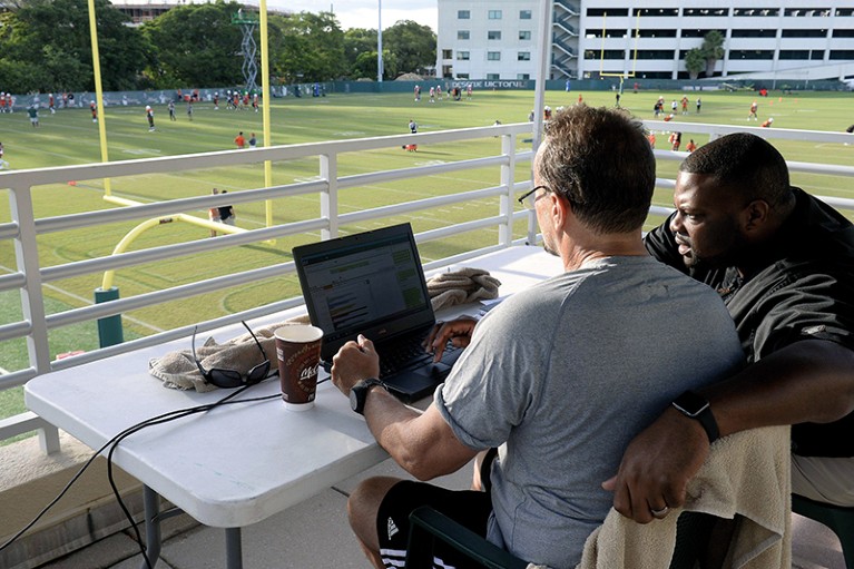 A coach and data analyst sit in front of a laptop overlooking a football pitch