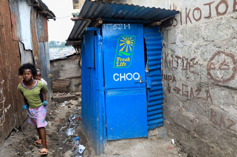 A woman carries a child on her back as she walks by a makeshift toilet booth in the Mathare slums, Kenya