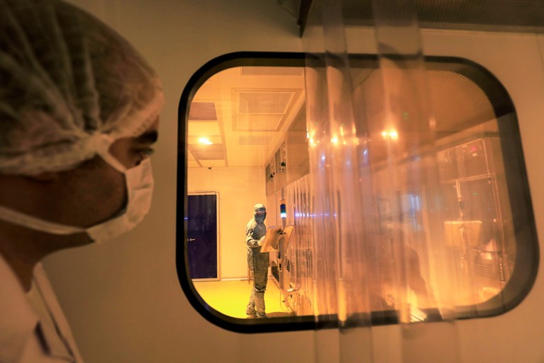 一名戴着口罩的男子透过窗户向制药实验室望去，旁边是一名穿着个人防护装备的实验室技术人员