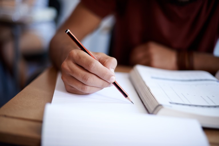 A person holds a pencil over a piece of paper while reading from an open book