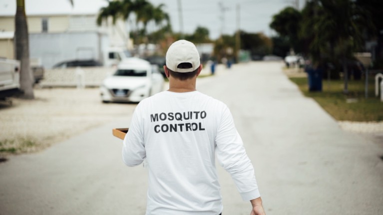 A man walks down a road in Florida holding a tray and wearing a shirt that reads 'Mosquito Control' on the back