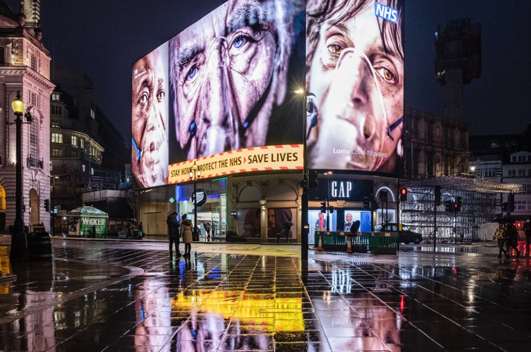 An electronic billboard at night in London shows three images of elderly people wearing oxygen masks and 'stay at home' messages
