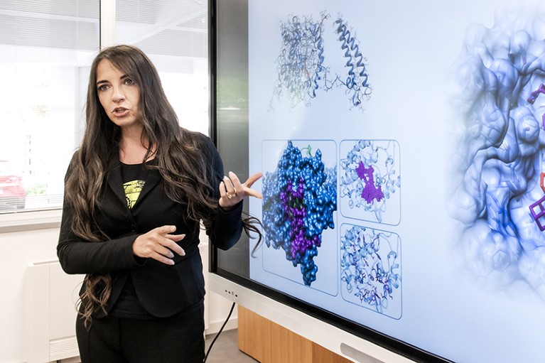 Lidia Pieri stands in front of a meeting room screen pointing at a digital COVID-19 structure