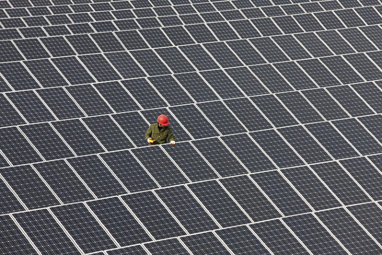 A figure in a hard hat and high visibility gear examines a solar panel in the centre of a large solar array