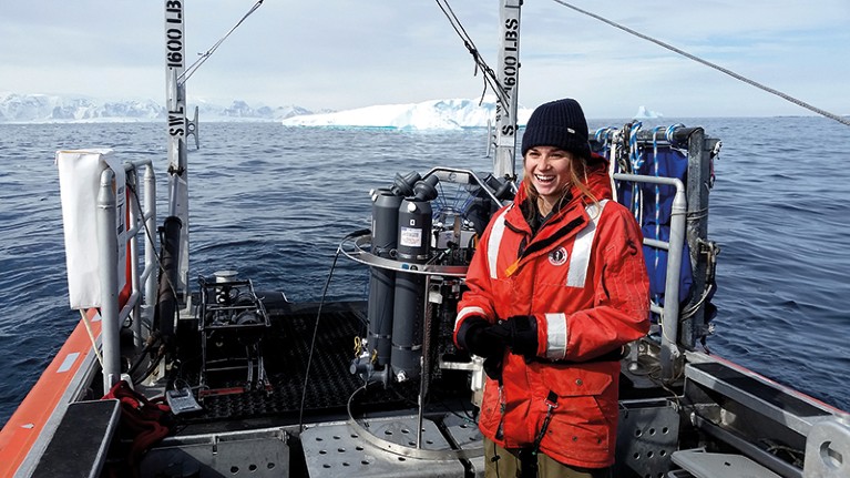 Anna Wright stands on the deck of a ship in winter gear with Antarctica in the distance