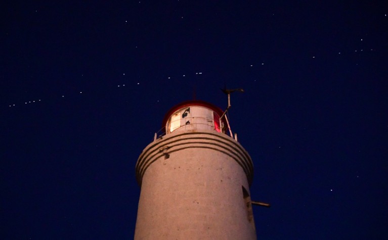 A string of the Starlink satellites streak diagonally across the sky above a lighthouse at night