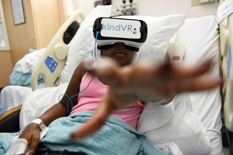 A young girl reaches towards the direction of the camera while wearing a VR headset in a hospital bed