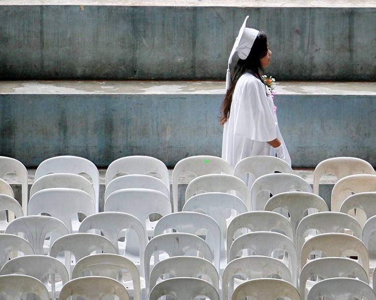 A graduate in a white robe walks by rows of empty chairs.