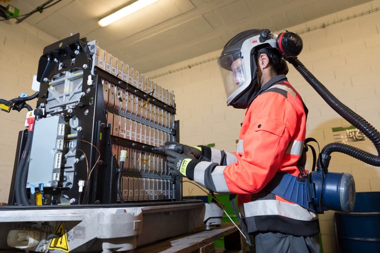 A worker wearing a ventilated gas mask dismantles a electric vehicle battery with a hand drill