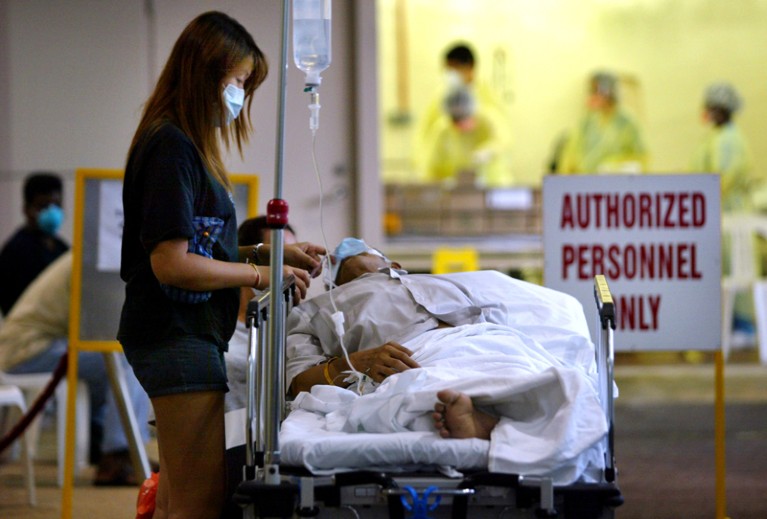 A woman wearing a face mask stands over a person in a hospital bed, with an intravenous drip.