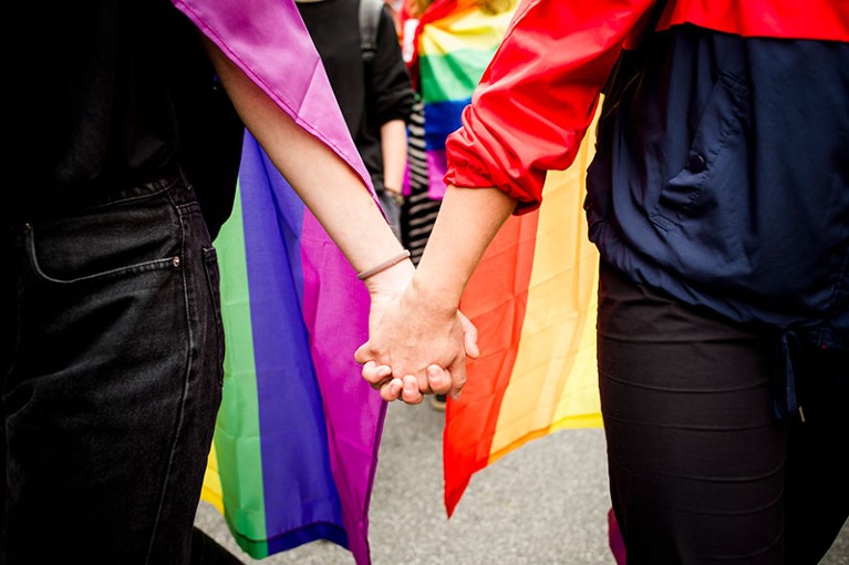 Two people hold hands while wearing LGBT+ rainbow flags during an anti-discrimination march.