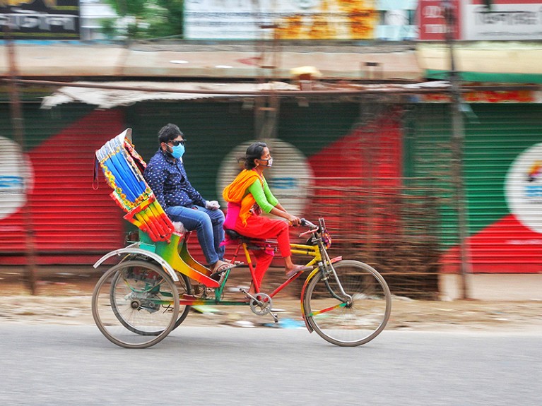 A woman wears a protective mask while driving a rickshaw with a masked passenger.