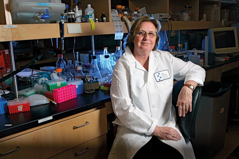 Wearing a lab coat, Jeanne Loring sits in front of a bench covered in lab equipment